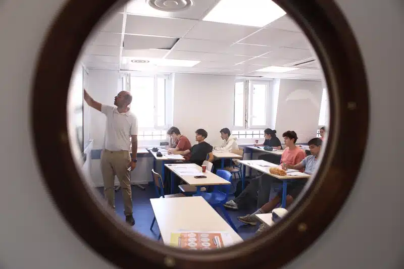 Salle de classe du stage intensif de la Toussaint de Groupe Réussite, vue à travers le hublot de la porte, avec un professeur au tableau et des lycéens au travail